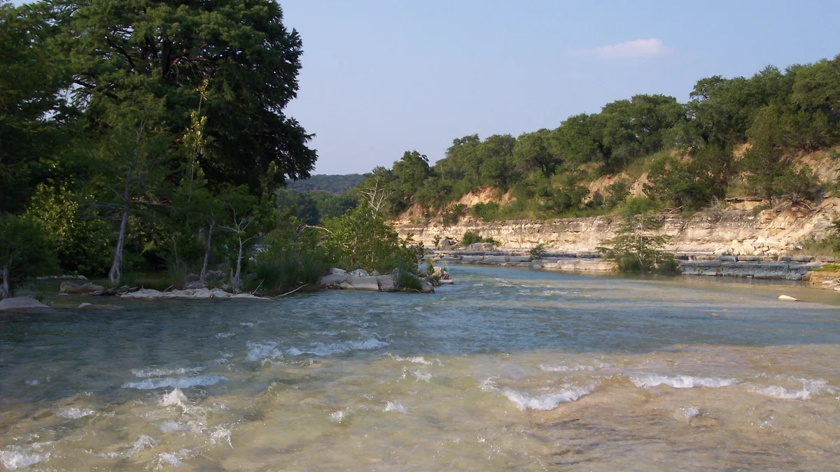 The Blanco River flows past trees and a limestone riverbank in Hays County, Texas.