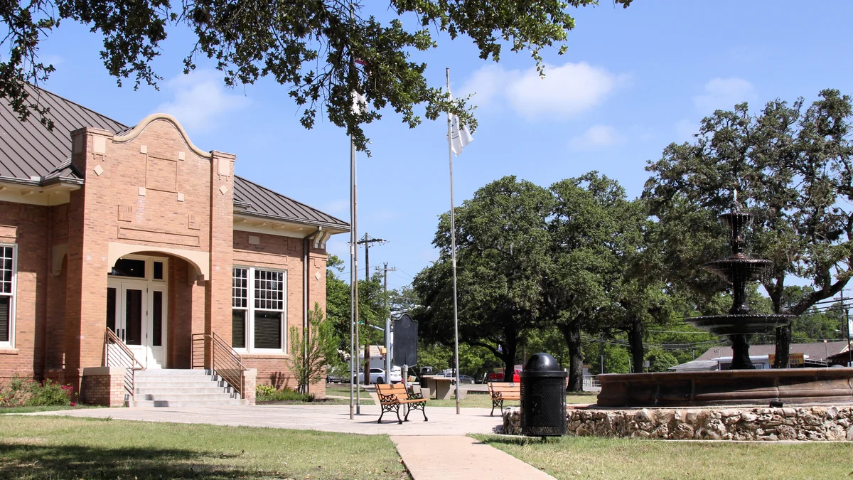 A fountain, shaded lawn, and brick building at City Square Park in Kyle, Texas.