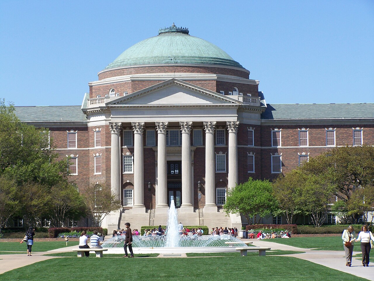 Dallas Hall at Southern Methodist University in Dallas, Texas — iconic Georgian red brick building with classical columns and a green copper dome, viewed from the campus lawn under a bright blue sky with students and fountain in the foreground. Independent campus housing information for SMU.