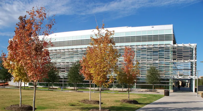 The Student Services Building at the University of Texas at Dallas in Richardson, Texas — a modern LEED Platinum campus building with mixed materials and glass, viewed from the campus walkway with mature trees and blue sky. Photo: Stan9999 / Wikimedia Commons (public domain). Independent campus housing information for UT Dallas.